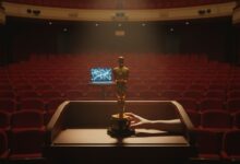 Golden Oscar statuette centered on wooden podium in empty theater with red seats, one hand on base, faint laptop glow in background.